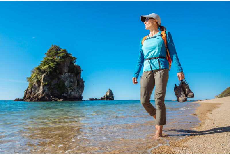 Woman wearing blue shirt walking on the beach holding her hiking shoes