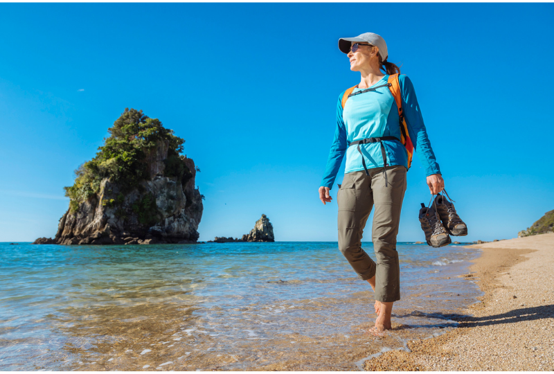 Woman wearing blue shirt walking on the beach holding her hiking shoes 