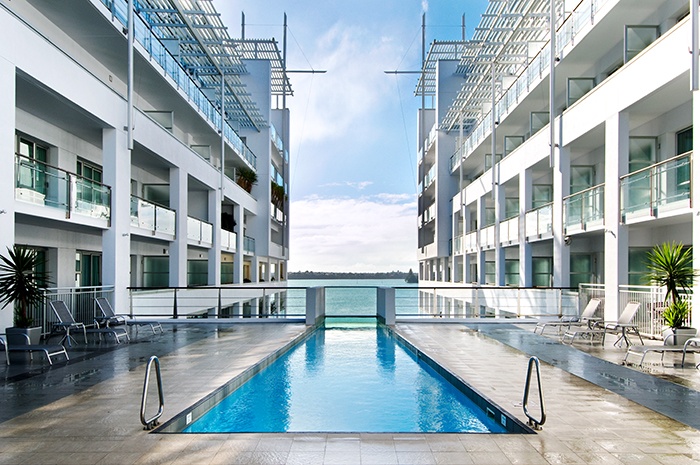 A long swimming pool located in the middle of two buildings with the sight of the sea