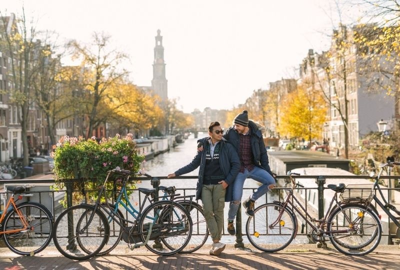 Two masculine presenting people stand in front of a canal in amsterdam with bikes next to them. One sits on a railing and has their arm around the other, they are smiling at each other