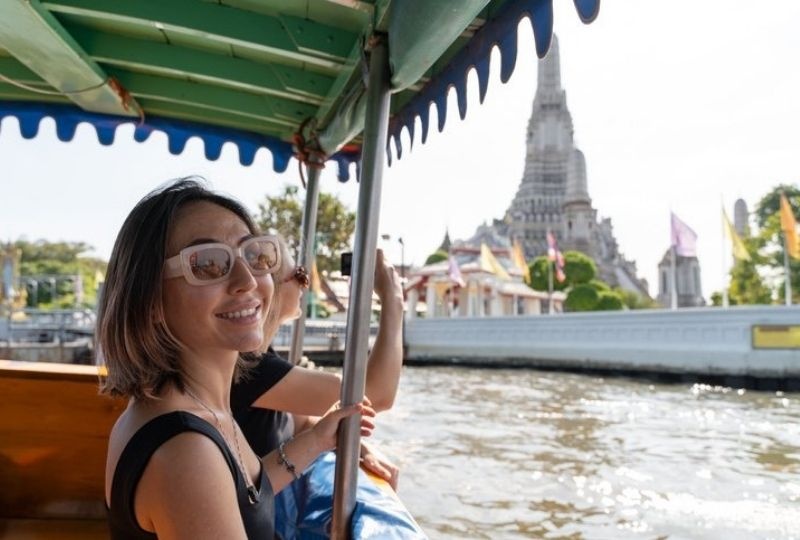 two femme presenting people sit in a boat, one is looking towards the camera with light pink glasses, smiling. The other holds a camera. There is a wat in background in bangkok