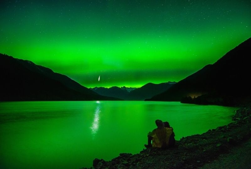 two people sit on the bank of a lake, one is wearing a hat. The image has a green tinge due to the northern lights 