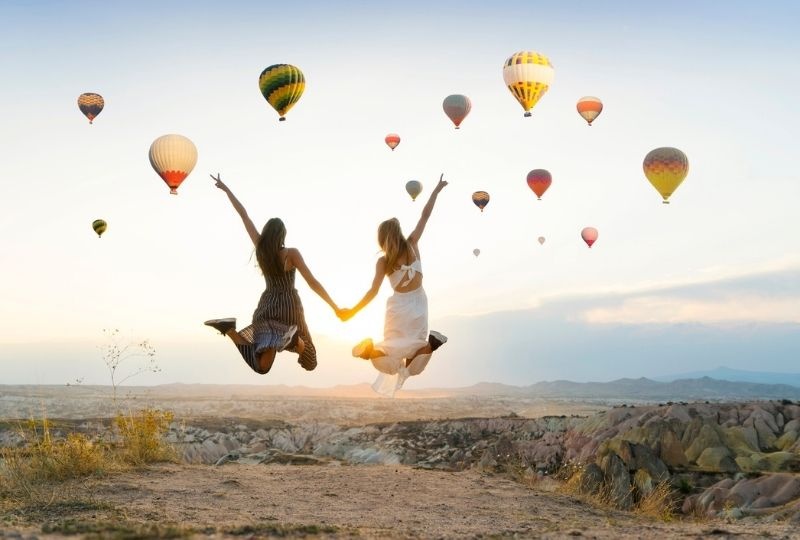 two femme presenting people in dresses jump into the air holding hands with other arms raised in air doing peace signs. Iconic hot air balloons are in the air as the sun rises
