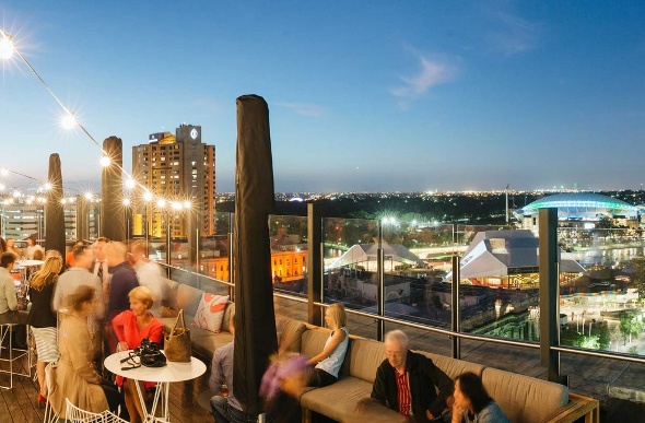  a well-lit rooftop restaurant overlooking the Adelaide city and full of happy people sitting on couches