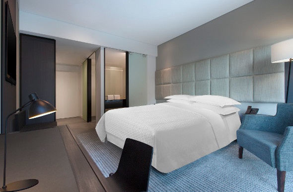  a minimalist hotel room with a clean white king-sized bed, blue bedside chair,  light blue carpet, and black lamp