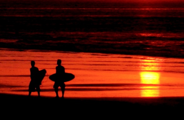  a silhouette of two men carrying their surfboard across the shore during late sunset