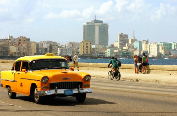  a taxi passing along the Cuba Havana Malecon with people overlooking the sea and a person riding a bike