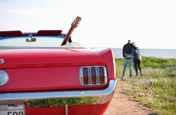  a red covertiblle car with a guitar inside and a couple hugging each other on the background