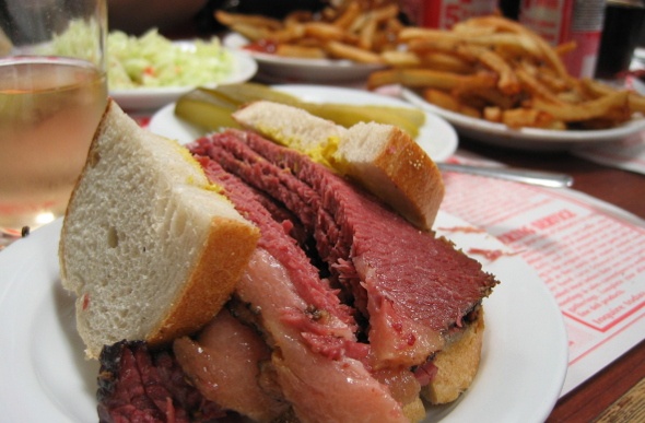 A sandwich loaded with corn beef and bowls of chips in the background