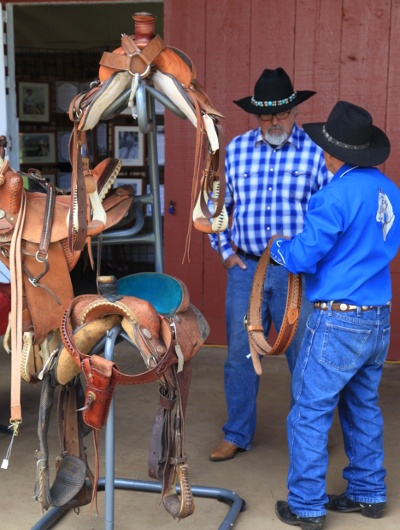  Two men in blue shirts and black cowboy hats next to horse saddles 
