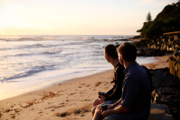  two men sitting on beach watching sunrise at moffat beach