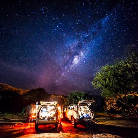 Cars parked in the woods beneath a star-studded purple sky