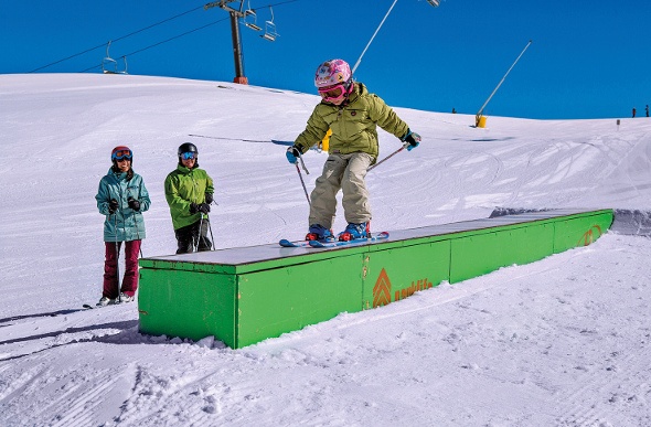 a kid practicing to ski on a green platform with his parents cheering for him on the  side