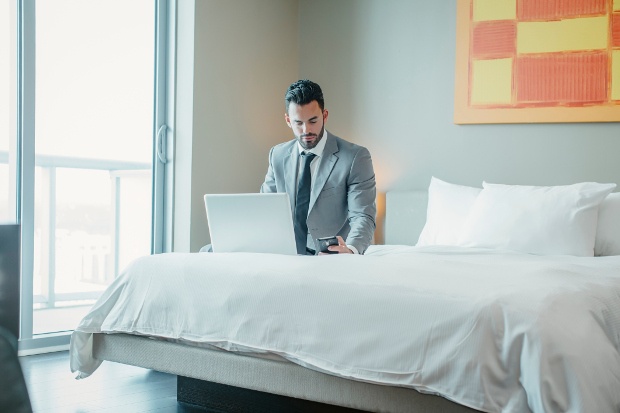 A man sitting on a hotel bed checking his phone with his laptop open