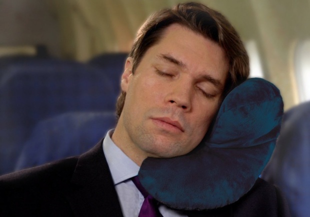 Man wearing a suite, asleep on a plane with a blue travel neck pillow on his shoulder