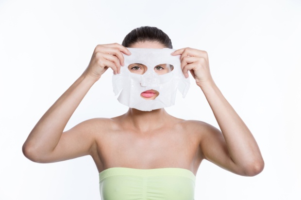 A woman wearing a towel and holding a face mask, with cut outs for her eyes and mouth, in front of her face