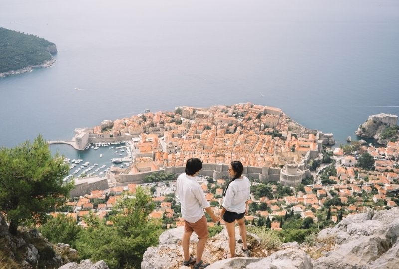  One masculine and one femme presenting person of asian background stand on a cliff top with the city of Dubrovnik