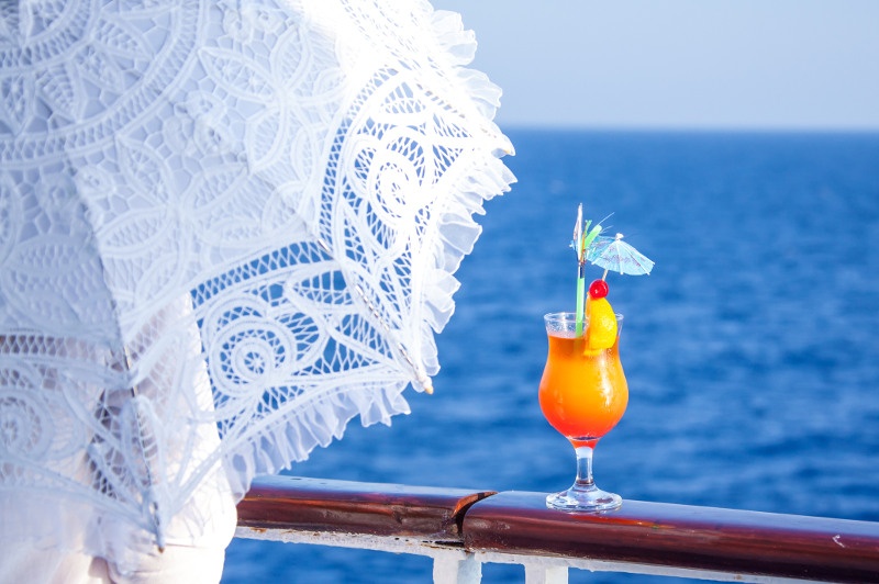 A woman with a parasol rests her drink on a cruise ship's railing.