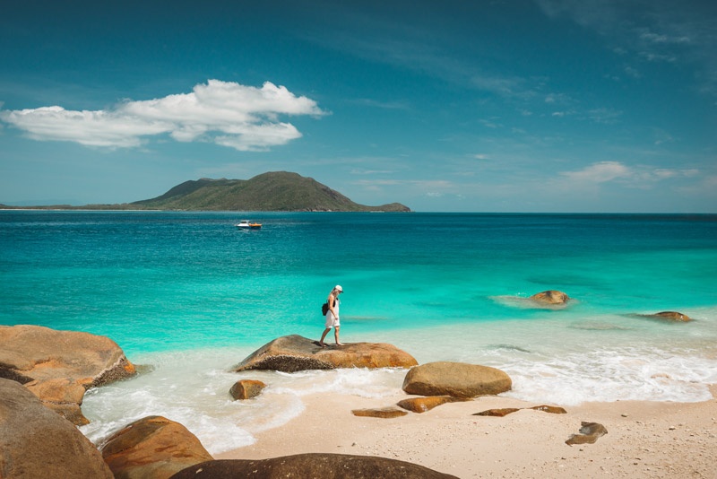 Woman standing on a rock on a white sand beach with clear blue water and mountain views 