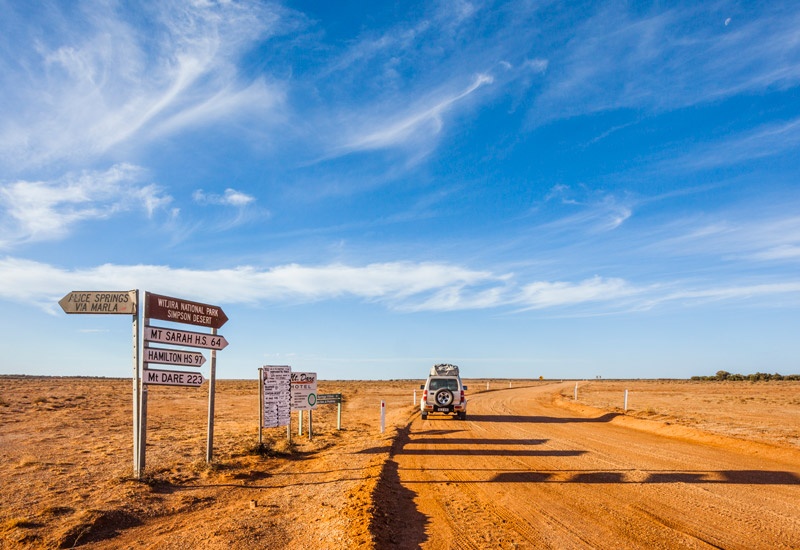 A silver car driving on a red dirt road next to various street signs 