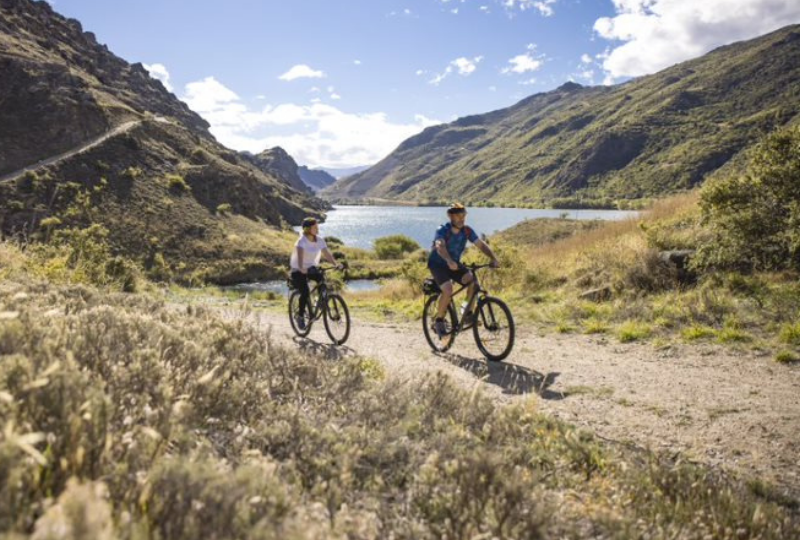 a couple bike riding next to Lake Dunstan on a sunny day 