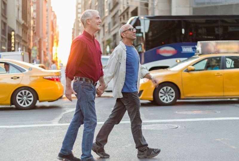 two older masculine presenting people walk holding hands. Yellow taxis are behind the, and they are in New York City 