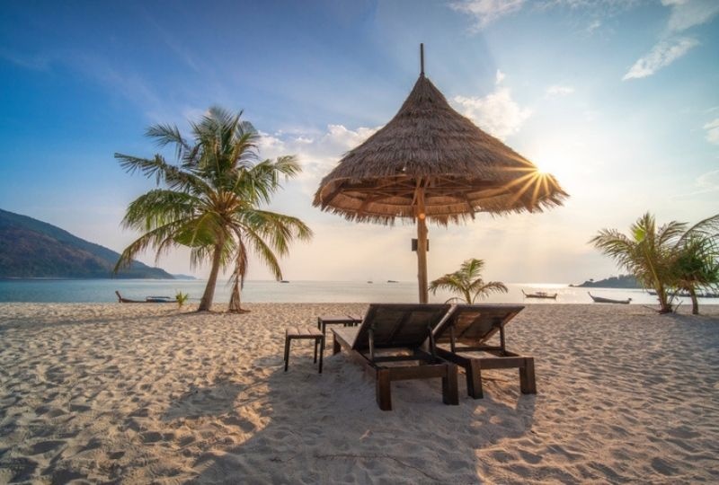 two wooden pool lounge seats under a nipa umbrella surrounded by palm trees in the shore of Phuket beach