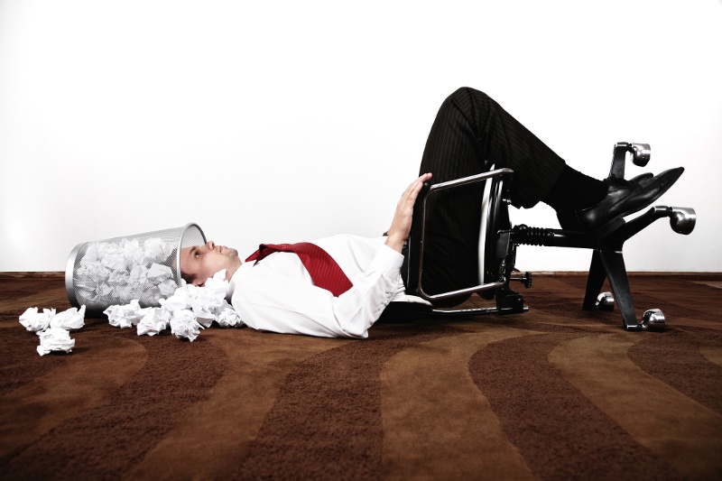 A businessman lies on his back with his head in a waste paper basket.