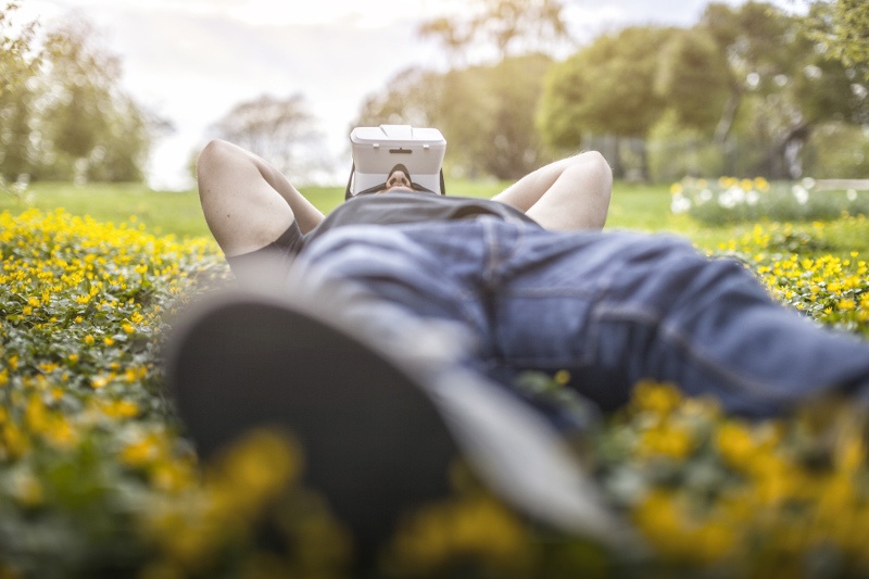 A man wearing virtual reality goggles lies in a field of yellow flowers.