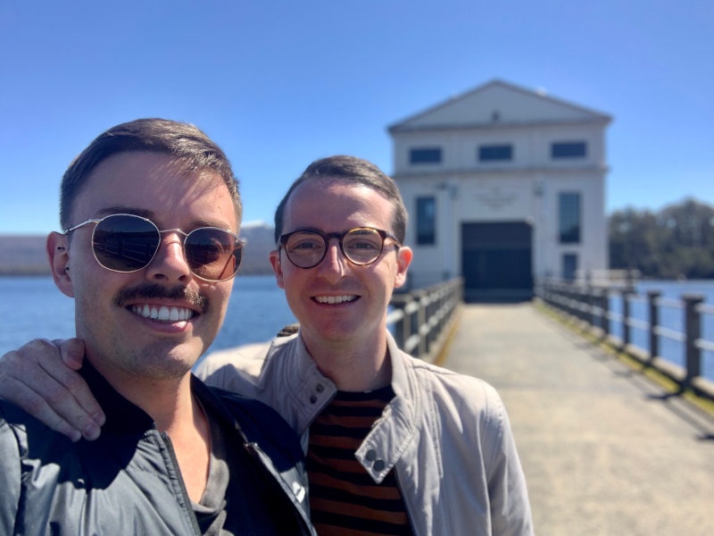 two men smiling and taking a selfie in front of the pumphouse 