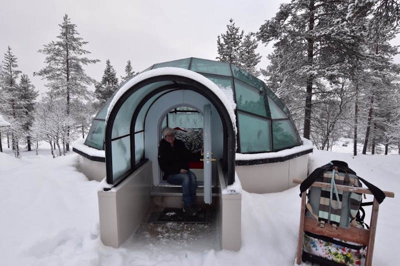 Michelle rugged up holding the door open of a glass igloo. 