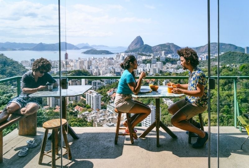 two people sit at a small table high up on sugarloaf mountain with plates and cups in front of them. one is masc presenting and the other is femme presenting. Another masc presenting person sits alone with legs up on seat. Rio is in the background 