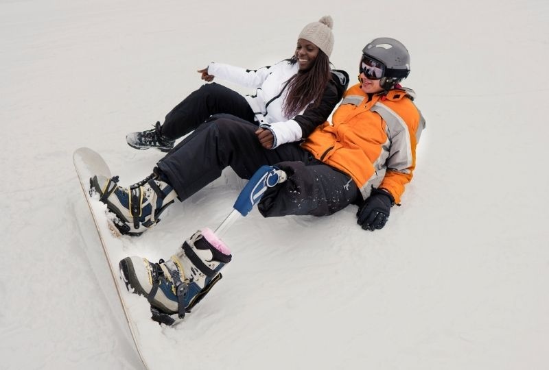 one black femme presenting person and one white masc presenting person with prosthetic leg are on their back in snow with snowboard and ski gear