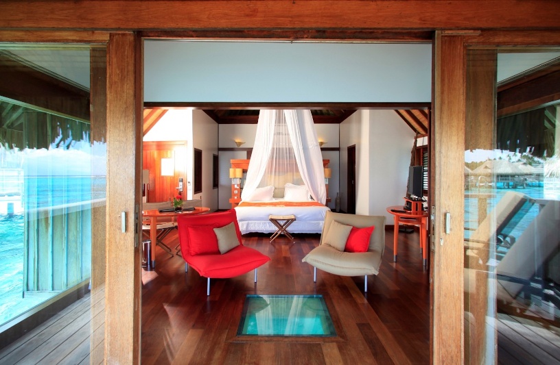looking into an overwater villa and into the main bedroom. There is a small glass bottom built into the floor