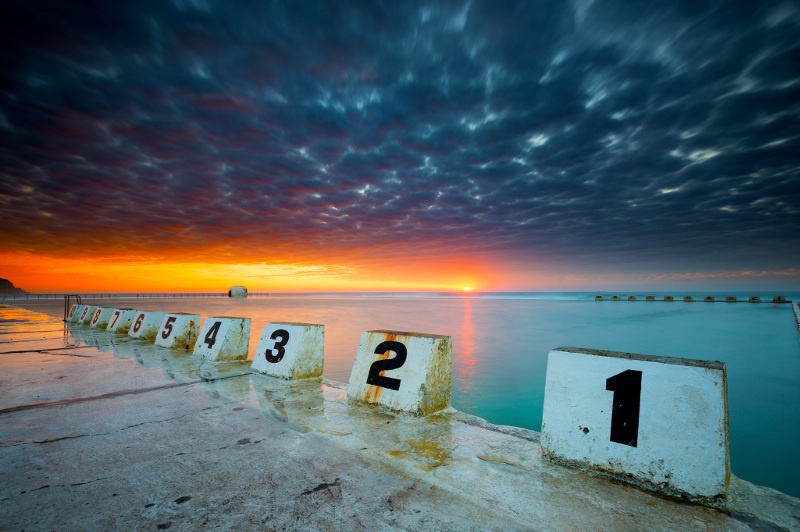A low-angle view from the corner of Newcastle's Merewether Ocean Baths, as the sun rises over the sea in the background.