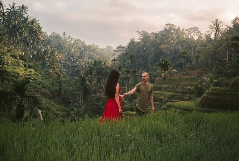 A femme presenting person wearing a bright red long dress holds the hand of a male presenting person wearing a green shirt. They are in a rice paddy field in Ubud, Bali.