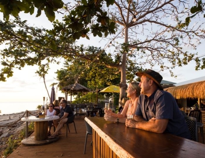 Man and women sit at outdoor bar having a drink and smiling. Other people sit at other tables in the background