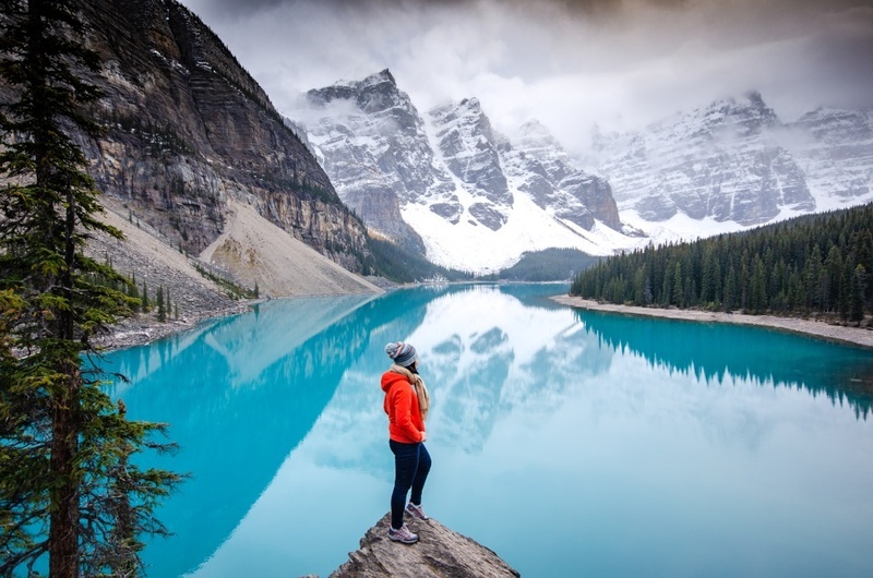 A lady standing at the edge of a stone looking at the sea that surrounded by mountains and snow