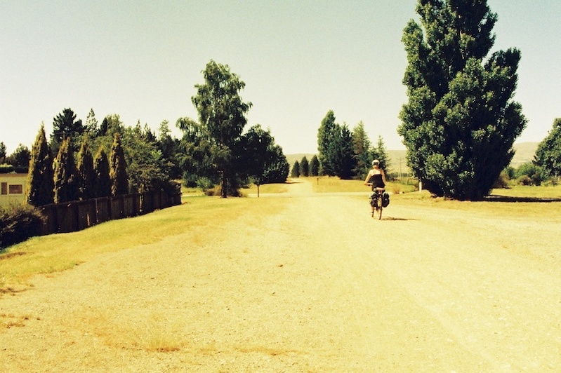 A woman rides a bike along a broad dusty trail with trees here and there on each side.