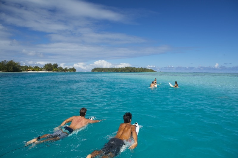 Two men paddle out on surfboards to a group of friends gathered in the water with tropical islands in the background.
