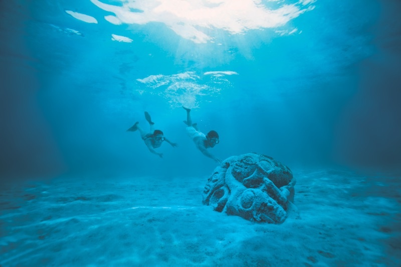 An under water shot of two divers in masks swimming towards a large submerged stone carving.