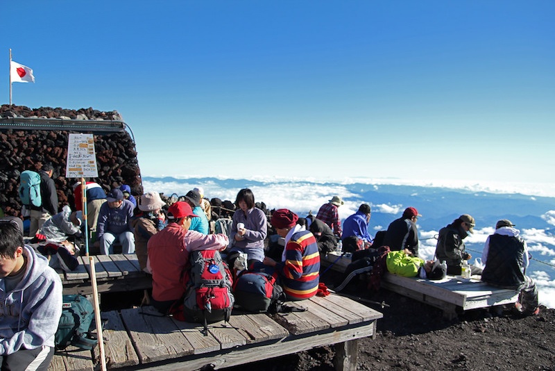 Climbers sit on wooden benches on the summit of Mt Fuji on a clear day.