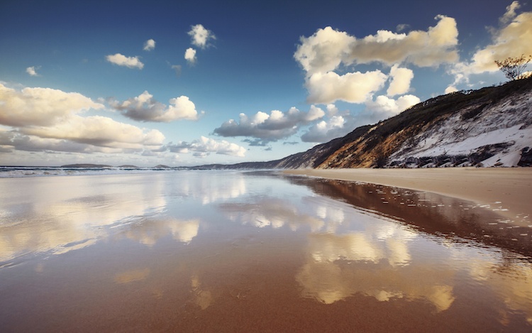 Clouds in a blue sky are reflected in still shallow on a wide beach by sand cliffs at Rainbow Beach, Queensland