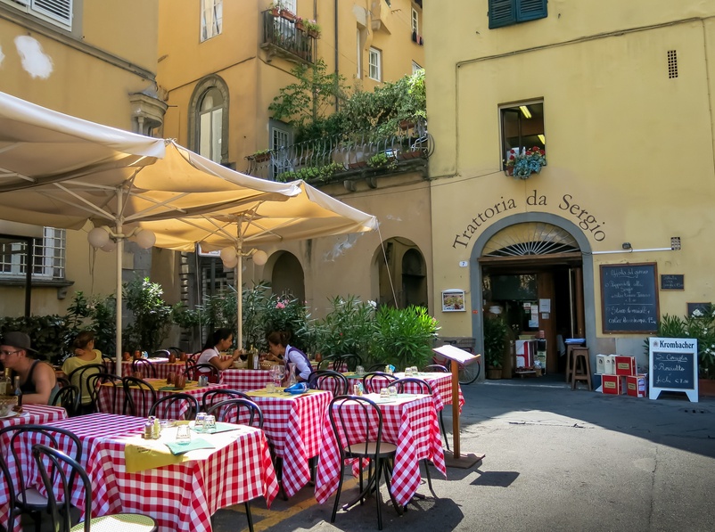 People sit at tables with red-and-white checked tablecloths at an el fresco cafe in front of a large Romanesque building with tall archways.