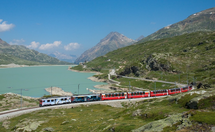 A passenger train winds its way around a lake in a green mountainous region of Switzerland