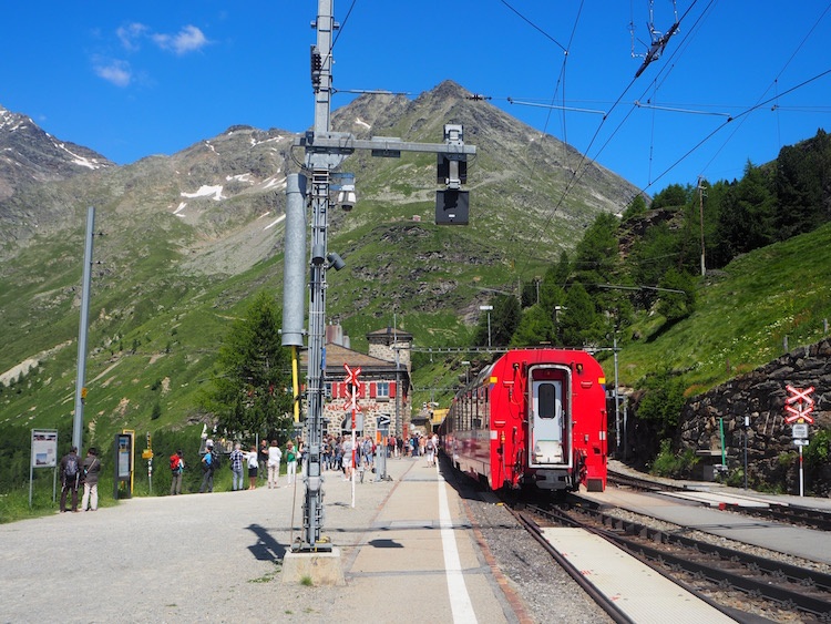 A red passenger train waits at a station with large mountains in the background on a clear day in Switzerland.