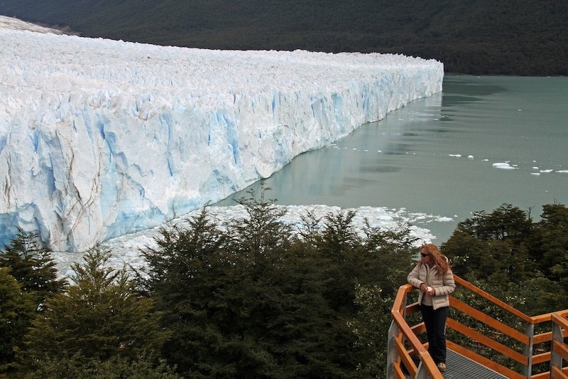 A woman on a lookout platform looks out over  the vertical wall of a huge glacier sprawling over a lake