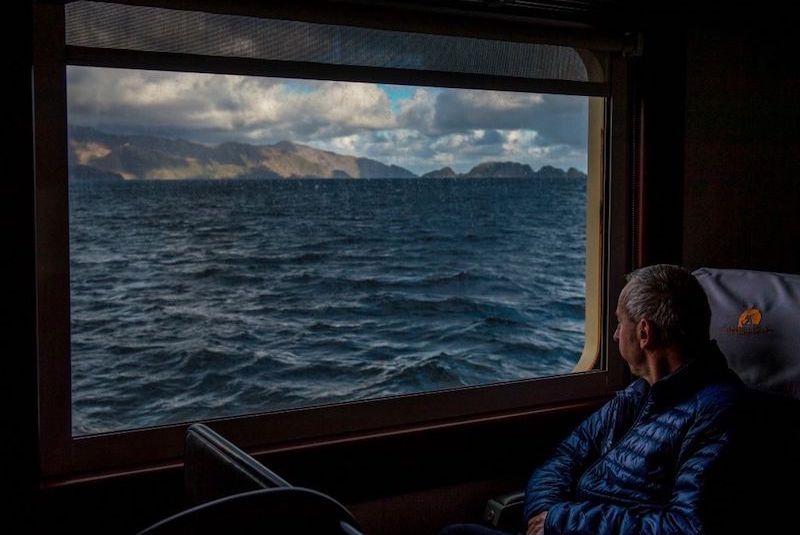 An elderly man looks out a window over a choppy sea and landforms on the horizon