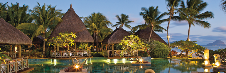 Shelters with thatched rooves among palm trees by a swimming pool in the early evening