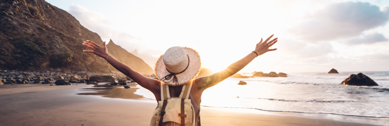 female presenting person wearing a straw hat and yellow shirt stands on beach with arms wide open